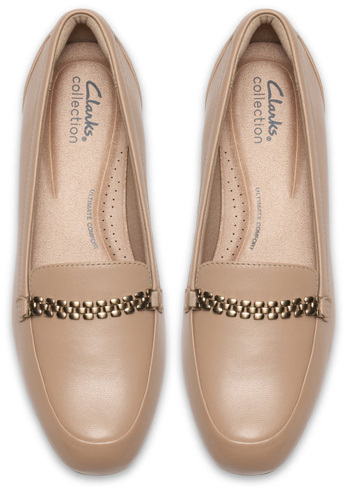 A pair of nude-colored, leather slip-on loafers, viewed from a top-down angle, featuring a metallic chain-link detail across the instep and 'Clarks Collection' branding printed on the cushioned insole.