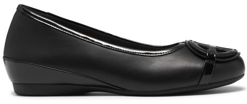 A black slip-on flat shoe with a low wedge heel, featuring a smooth faux leather upper and a decorative circular emblem on the rounded toe.