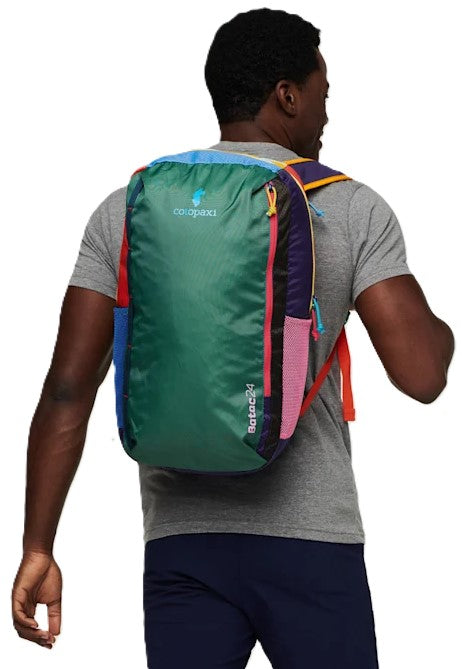 A person viewed from behind wearing a multi-colored backpack with a large green panel, pink side, blue accent, orange straps, and a zipper with red trim, featuring text that reads 'cotopaxi' and 'Bataac24'.