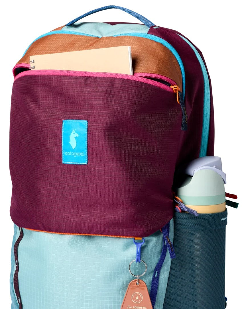 A brightly colored, multi-panel backpack featuring a maroon main compartment with a light blue label bearing a llama icon, a light blue lower section, tan accent trim, and a side pocket holding a teal reusable water bottle.