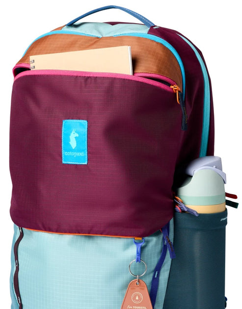 A brightly colored, multi-panel backpack featuring a maroon main compartment with a light blue label bearing a llama icon, a light blue lower section, tan accent trim, and a side pocket holding a teal reusable water bottle.