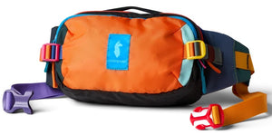 A colorful fanny pack with an orange front panel featuring a blue rectangular label with a white llama graphic and the brand name Cotopaxi, black sides and bottom, teal accents, and a multicolor adjustable strap with a purple buckle on one side and a red and yellow buckle on the other.