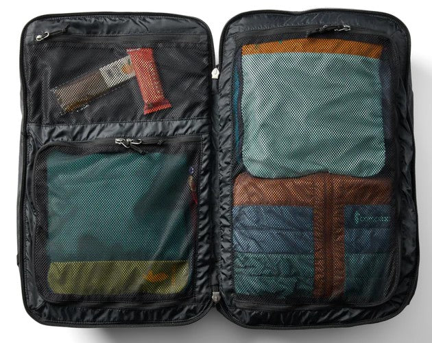 An open black travel bag interior showing multiple zippered mesh compartments filled with packed clothing and a small snack bar.