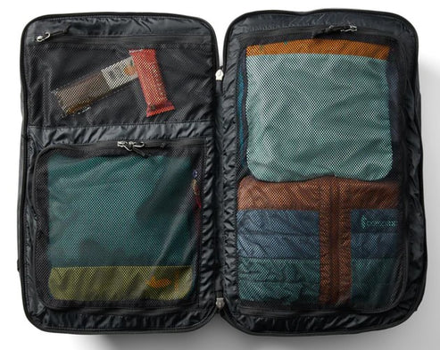An open black travel bag interior showing multiple zippered mesh compartments filled with packed clothing and a small snack bar.