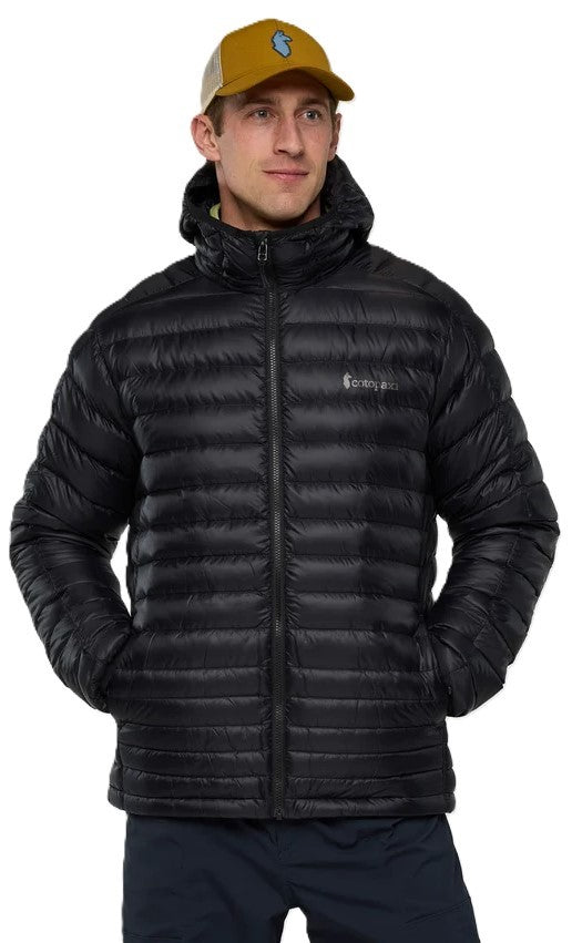 A man wearing a black quilted down jacket with a hood and a tan baseball cap with a blue circular patch, looking forward against a white background.