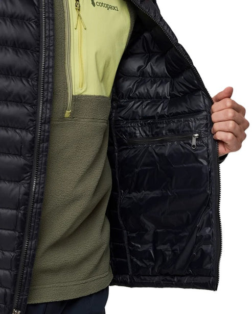 Close-up view of the interior of a black quilted puffer jacket, showing a zippered chest pocket and a hand reaching into the side, layered over a lime green and olive fleece top with visible Cotopaxi branding.