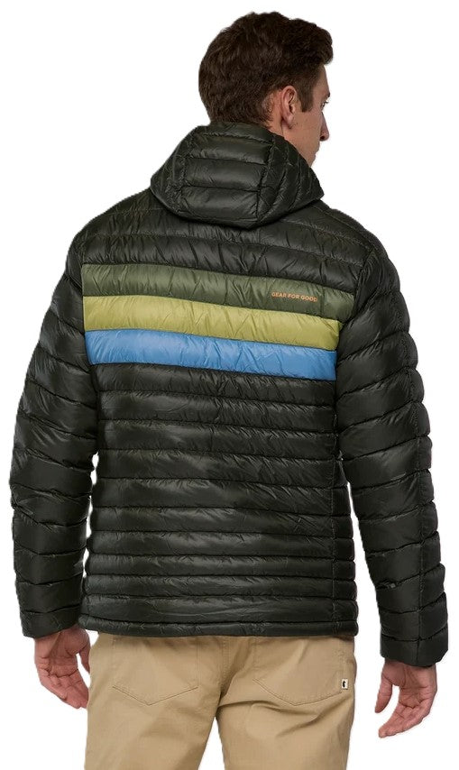 Rear view of a man wearing a dark olive green quilted hooded puffer jacket with a horizontal lime green and blue stripe across the upper back and light tan trousers.