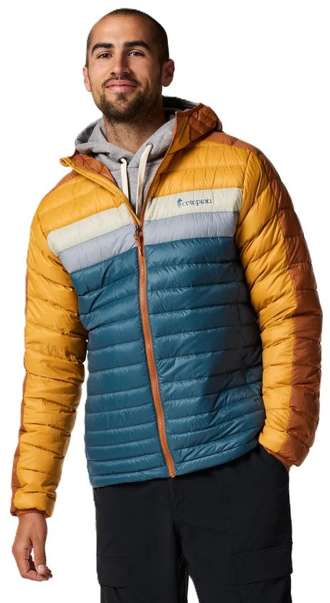 A man wearing a hooded, color-blocked, quilted down puffer jacket with yellow sleeves and hood, a horizontal grey stripe across the chest, and a teal body, layered over a light grey hoodie, paired with black pants.
