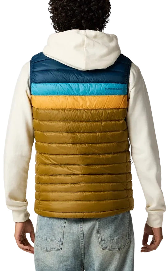 A back view of a person wearing a puffer vest featuring horizontal color-blocked sections in dark blue, light blue, bright yellow, and olive green over a cream-colored hooded sweatshirt and light blue denim jeans.