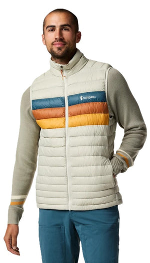 A man wearing a light beige, quilted puffer vest with three horizontal, multi-colored stripes of blue, rust, and yellow across the chest, paired with a long-sleeved, sage-green sweater and blue trousers.