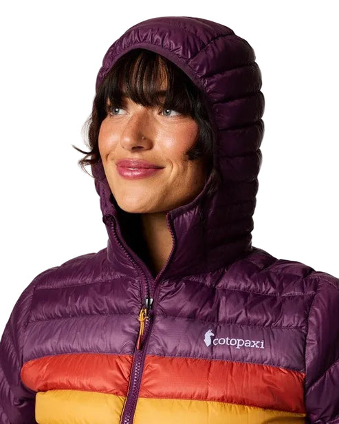 A woman wearing a color-blocked, hooded puffer jacket with dark purple at the top and hood, followed by a band of red and a band of yellow, featuring the word 'cotopaxi' printed on the chest.