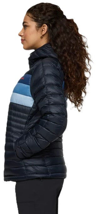 A woman seen from the side, wearing a hooded, quilted, dark navy blue puffer jacket with a horizontal light blue and blue stripe across the upper chest, paired with black trousers.
