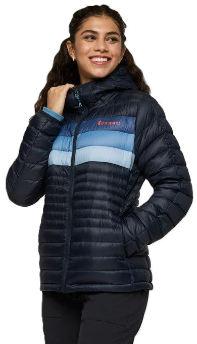 A smiling woman wearing a dark blue, hooded, quilted, insulated puffer jacket with a color-blocked design featuring horizontal bands of light, medium, and dark blue across the chest and the word Cotopaxi written on the upper chest.