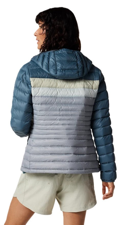 A back view of a woman wearing a color-blocked, hooded puffer jacket. The jacket features deep blue sleeves and a matching blue hood, transitioning into a light beige horizontal stripe on the upper back, followed by a series of grey horizontal quilted panels covering the rest of the back. She is wearing light-colored shorts.