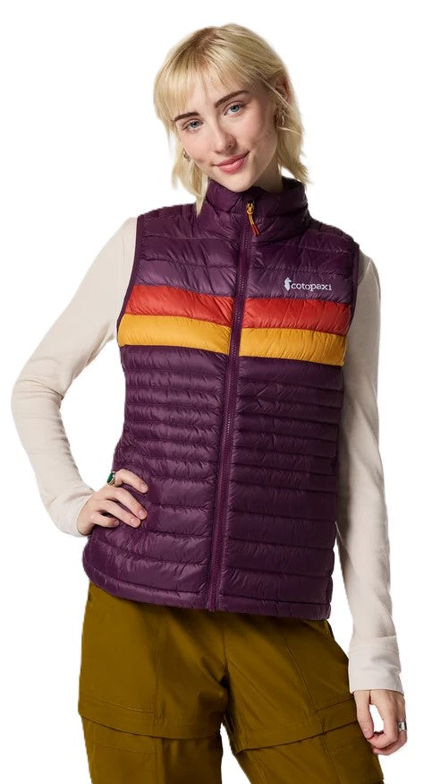 A woman wearing a dark purple, insulated quilted vest with a horizontal stripe across the chest featuring orange and yellow bands, over a cream-colored long-sleeved shirt and olive green pants.