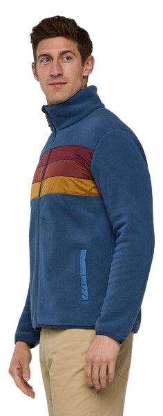 A man wearing a blue fleece jacket with a red, orange and yellow stripe across the chest and tan pants.