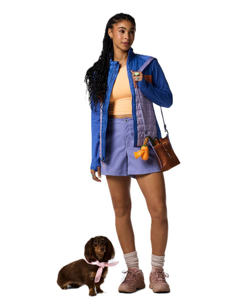 A woman wearing a blue jacket, orange top and lilac shorts stands with a dachshund wearing a pink ribbon. She carries a brown purse with charms, white socks, and pink athletic shoes.