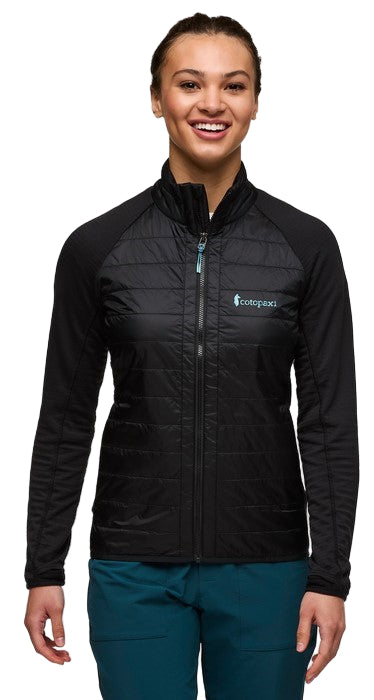 A woman wearing a black quilted jacket with black knit sleeves and dark teal pants, smiling.