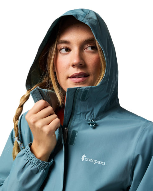Woman wearing a teal rain jacket with a hood; the logo 'cotopaxi' is visible on the chest.