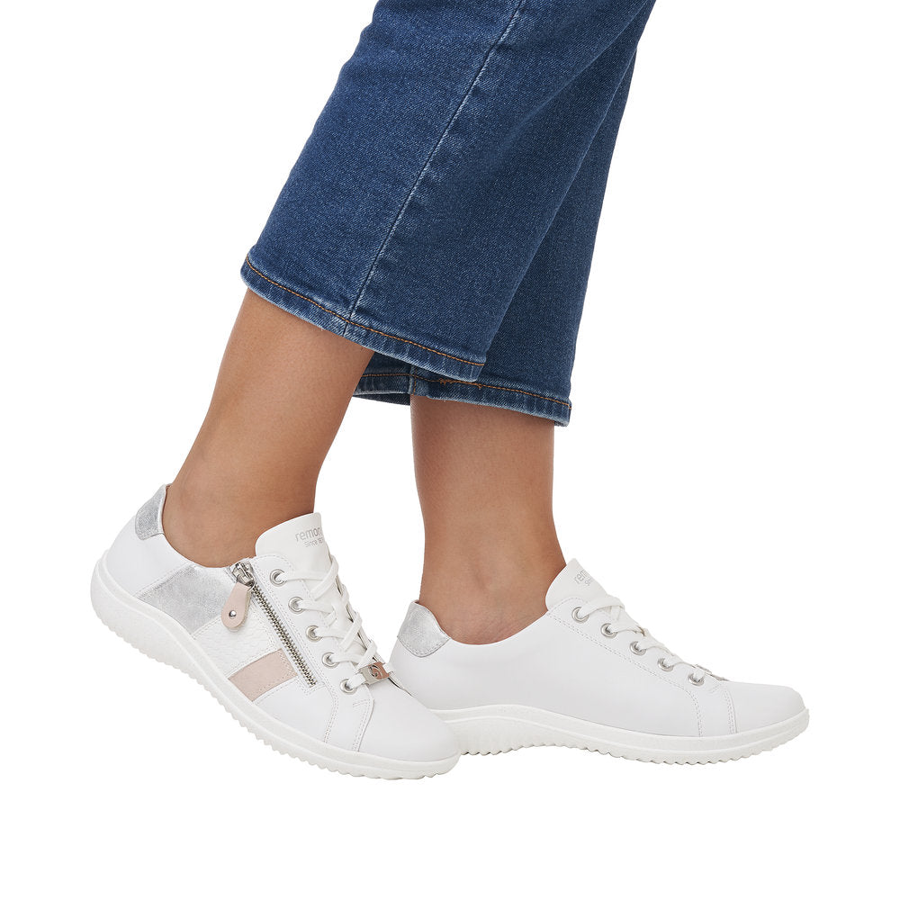 A person wearing white leather sneakers with a zipper on the left foot and silver and pink stripes.