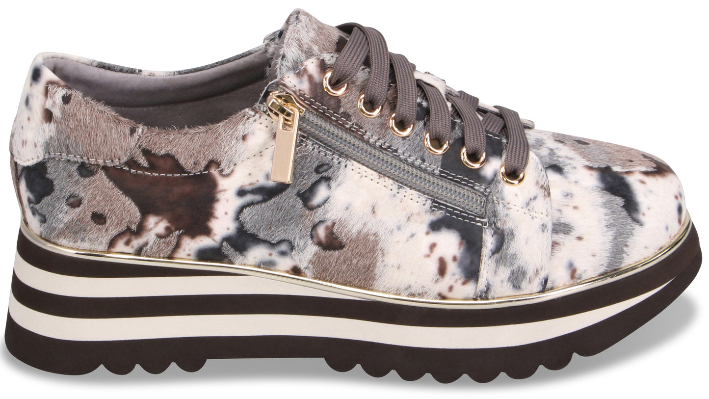 A low-top platform sneaker with a mottled, abstract black, gray, and white pattern, featuring a side zipper, gray laces, gold eyelets, and a thick striped black and white sole with a scalloped tread.