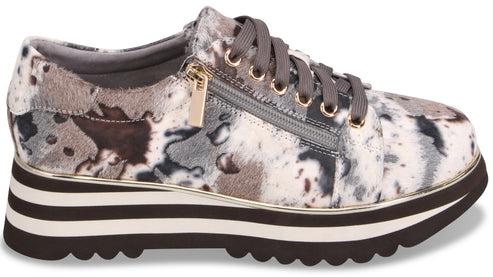 A low-top platform sneaker with a mottled, abstract black, gray, and white pattern, featuring a side zipper, gray laces, gold eyelets, and a thick striped black and white sole with a scalloped tread.