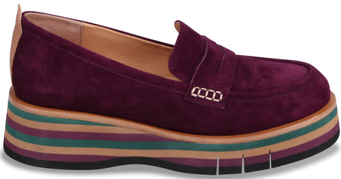 A plum-colored suede penny loafer featuring a stacked, multicolored striped platform sole with horizontal bands of tan, dark green, and purple, finished with a black rubber outsole.