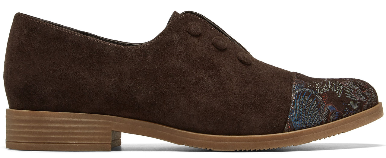 A brown suede slip-on shoe with a low, textured tan sole and a decorative, patterned dark blue and brown floral fabric panel at the toe.