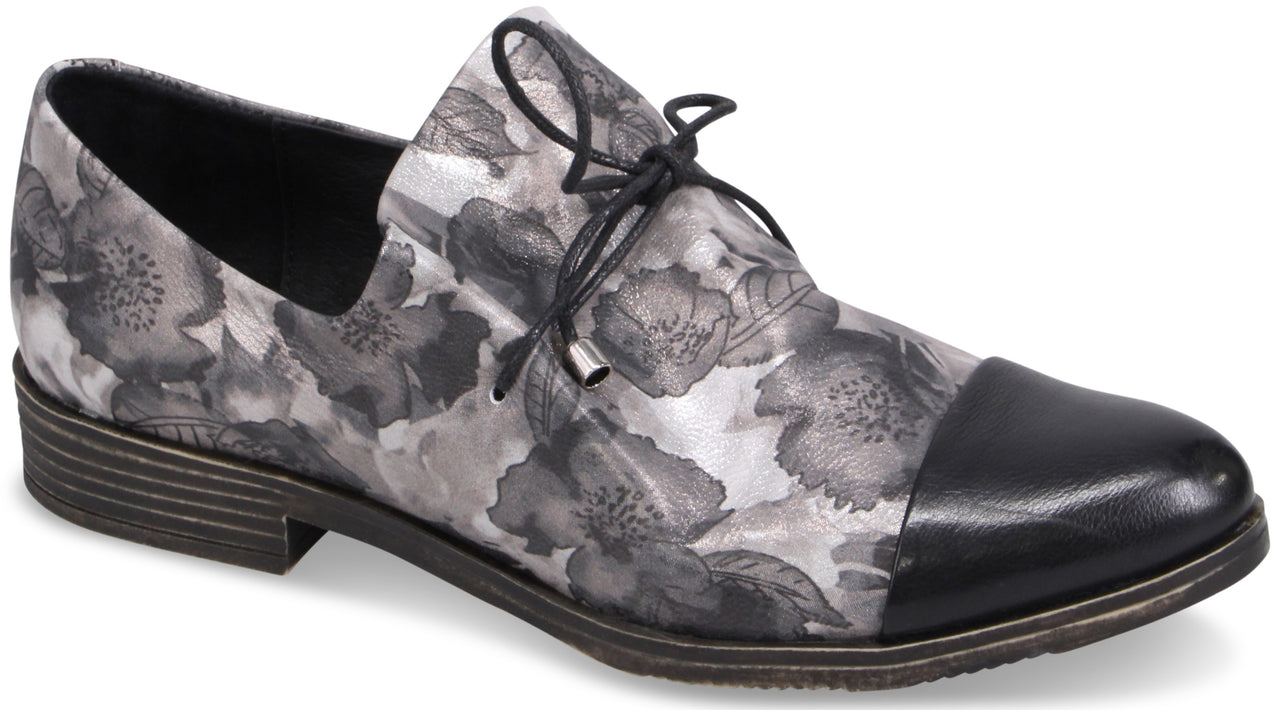 A women's flat lace-up oxford shoe with a black patent leather toe cap and a grey and black floral print body, featuring a dark brown sole and a small stacked heel.