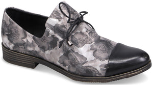 A women's flat lace-up oxford shoe with a black patent leather toe cap and a grey and black floral print body, featuring a dark brown sole and a small stacked heel.