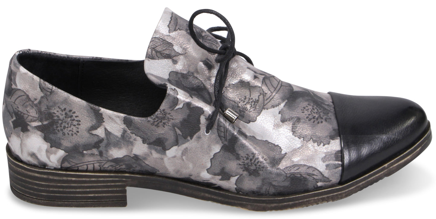 A women's flat lace-up oxford shoe featuring a grey and black floral print pattern on the body and a solid black smooth leather cap toe, finished with a dark low block heel.