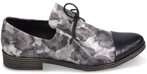 A women's flat lace-up oxford shoe featuring a grey and black floral print pattern on the body and a solid black smooth leather cap toe, finished with a dark low block heel.