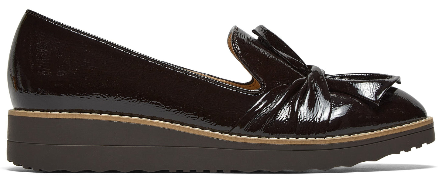 A dark brown patent leather slip-on wedge shoe with a decorative bow on the front, set on a stacked, serrated-tread platform sole.