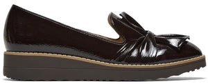 A dark brown patent leather slip-on wedge shoe with a decorative bow on the front, set on a stacked, serrated-tread platform sole.