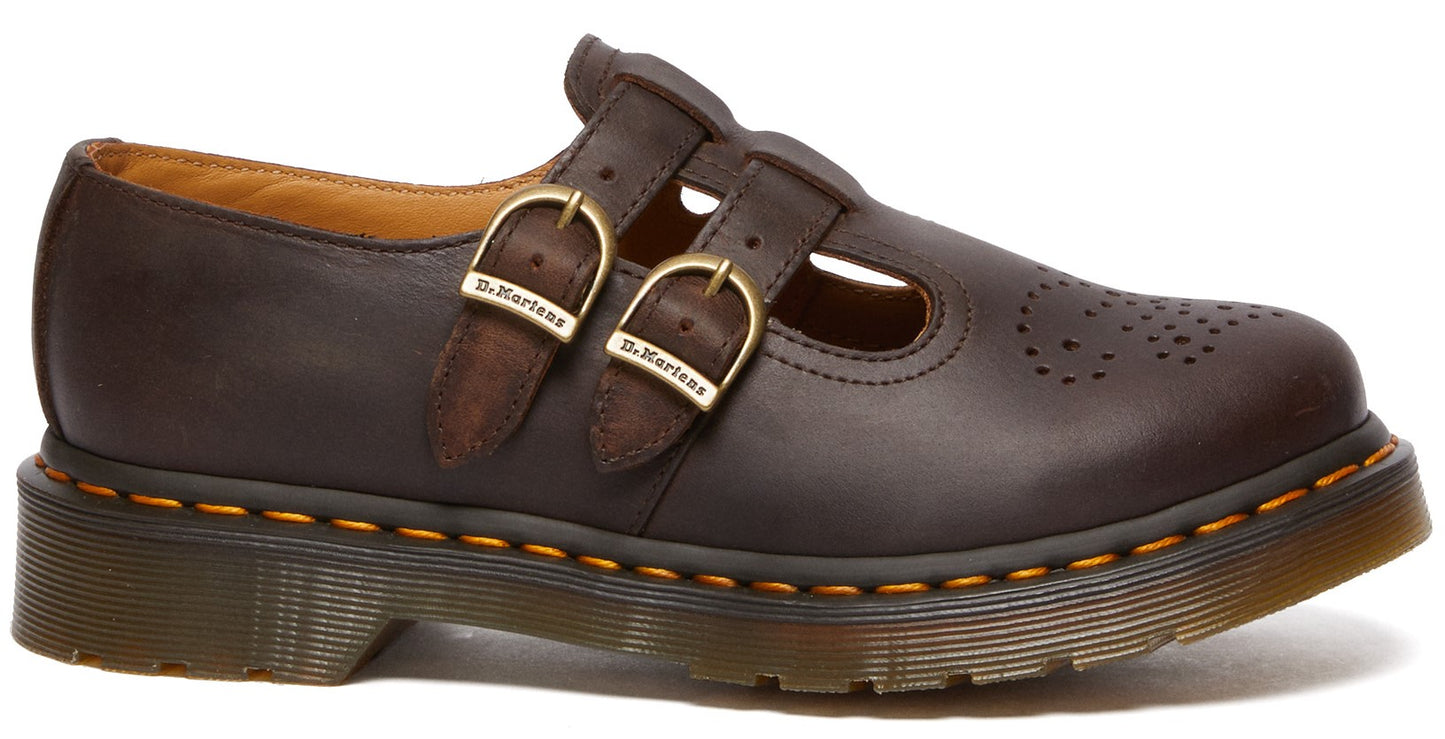 A dark brown leather Mary Jane shoe with two brass buckles featuring the text 'Dr. Martens', yellow stitching around the sole, and a decorative perforated pattern on the toe cap.