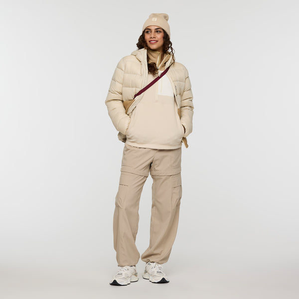 A woman wearing a beige beanie, cream puffer jacket, cream hoodie, beige pants and white sneakers, standing indoors.