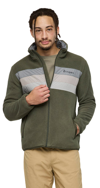 A person wearing a zipped, hooded, olive green fleece jacket with a gray stripe across the chest and khaki pants, standing.