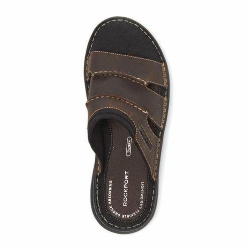 Top-down view of a brown leather slide sandal with black sole and white stitching, marked ROCKPORT and "shock absorbing".