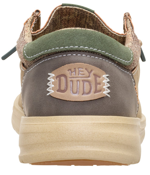 A rear-view close-up of a tan, olive green, and brown casual slip-on shoe with a tan platform sole and a patch on the heel that reads HEY DUDE in a stylized font.