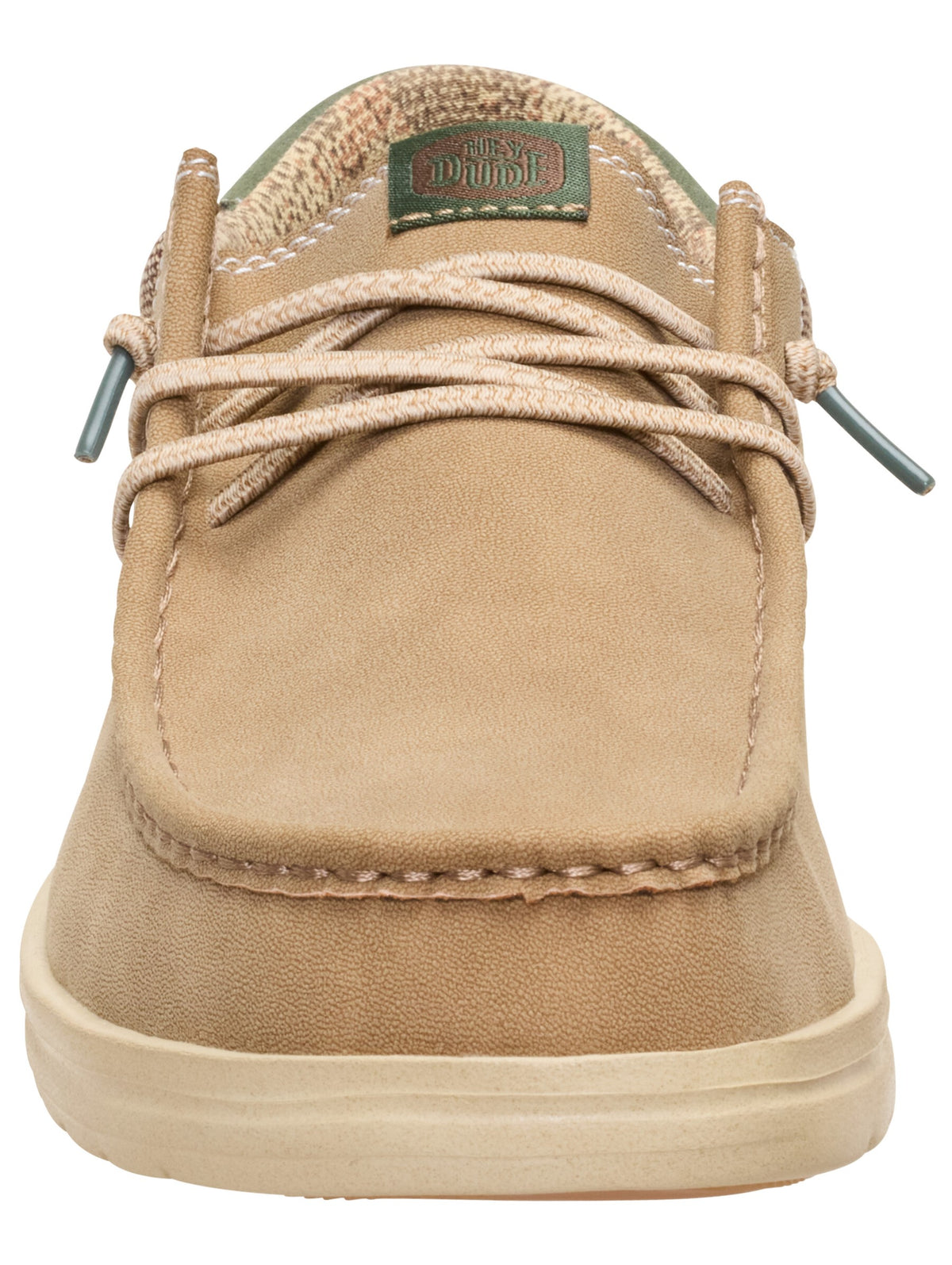 A tan slip-on shoe with a rounded toe, a visible stitched seam along the front, and beige laces in a crisscross pattern. A rectangular green tag with unreadable text is attached to the center of the tongue.