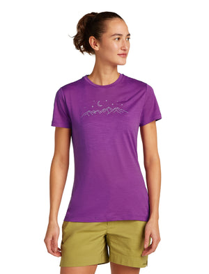 A woman wearing a purple T-shirt with a mountain line drawing and green shorts is standing.