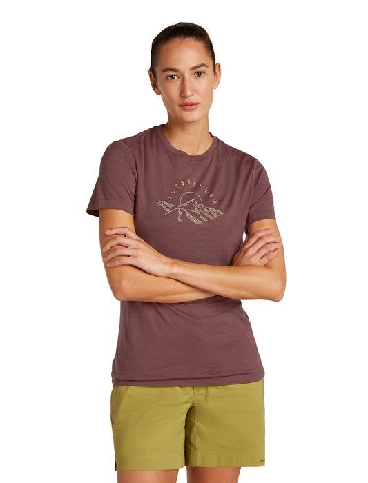 A woman wearing a brown t-shirt with a mountain and sun graphic stands with crossed arms over light green shorts.