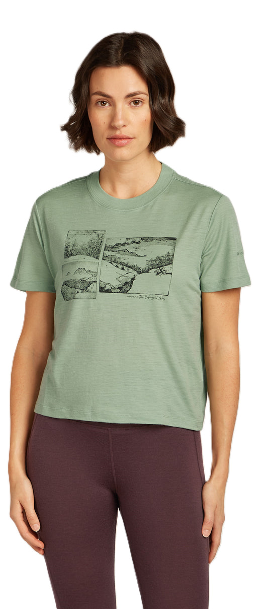 A woman is modeling a light-green, short-sleeved t-shirt with a black graphic of an outdoor landscape on the chest. She is wearing dark-purple leggings.