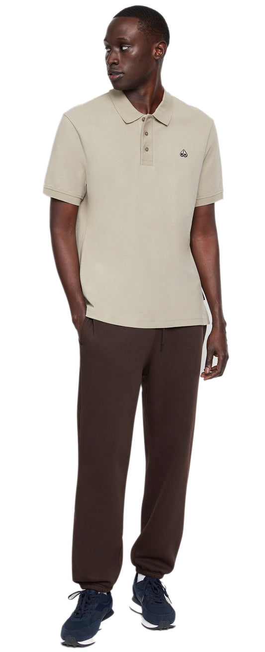 A man stands wearing a beige short-sleeve polo shirt and brown sweatpants with navy athletic shoes.