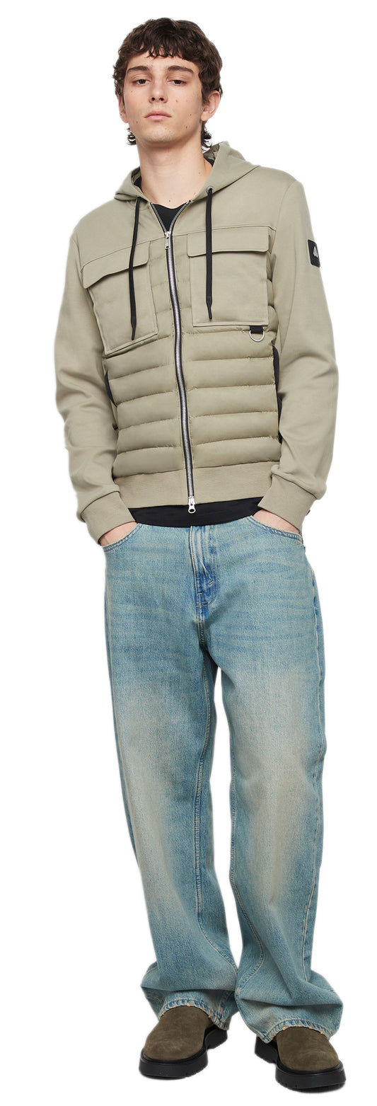 A person wearing a beige hooded jacket, a black shirt, baggy light blue jeans and brown platform shoes is standing and facing the camera.