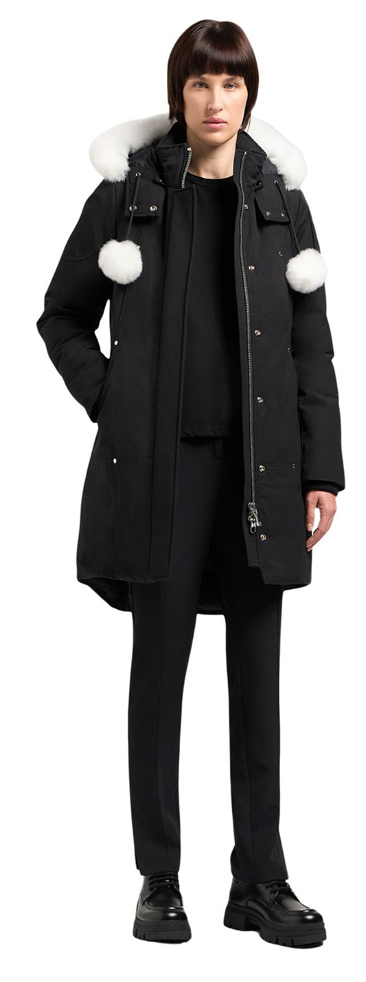 A person wearing a long black coat with white fur trim and white pom-poms, with black pants and black shoes.
