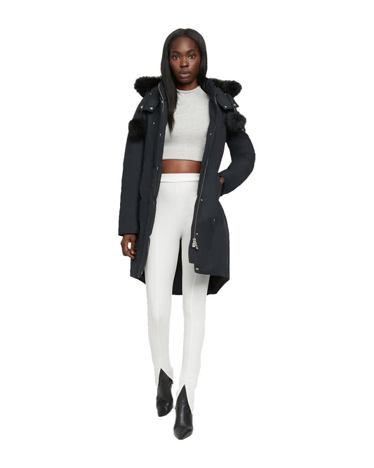A Black woman standing in front of a white background wearing a long black winter coat with a fur-lined hood, a white crop top and white pants.