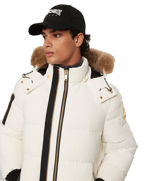 A person wearing a white puffer coat with fur around the hood and a black cap with embroidered white letters.