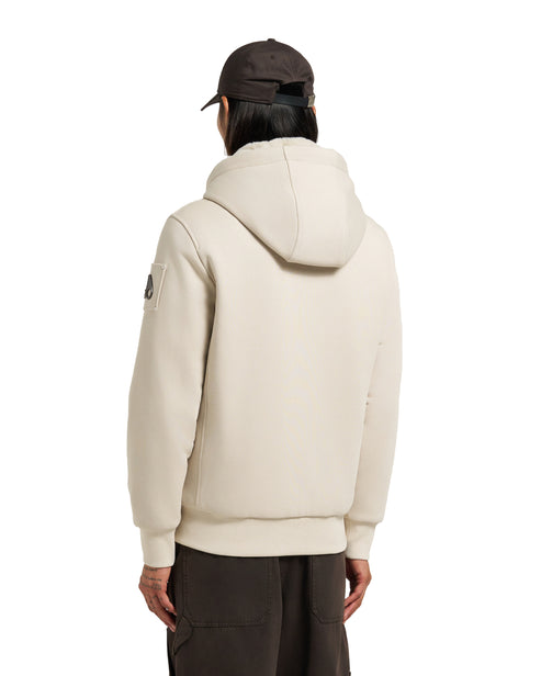 Man wearing a beige hoodie with a hood, facing away, with a patch on the left sleeve, and a brown cap.