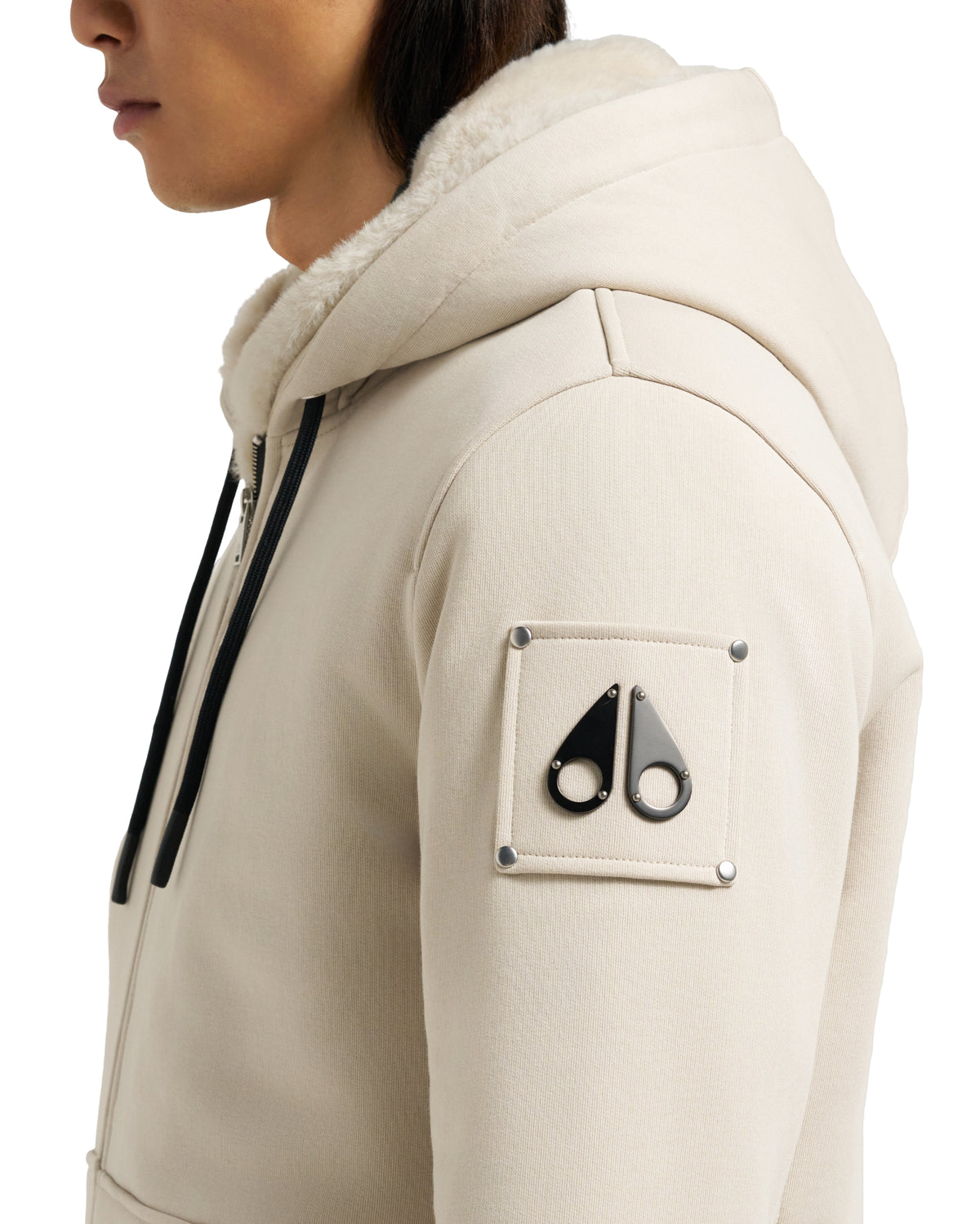 Close-up of a light beige jacket with fur-lined hood and a black moose knuckle logo on the left sleeve.